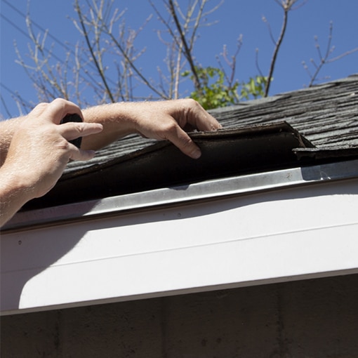 Hand lifting up roofing shingles to inspect storm damage