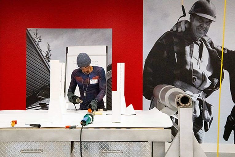 A GAF Roofing Academy student looking down while working in shop with an image of a GAF certified roofer behind him.